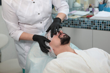 A beautician makes a mask on the forehead and around the eyes of a young man with a beard. Men's...