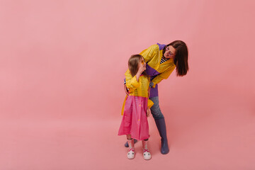 Lovely woman in trendy raincoat and jeans smiling and hugging her little daughter in stylish clothes on pink backdrop