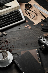 Male brutal dark gray desktop. On the table there is a typewriter, a cup of coffee and a camera, a notebook, sketches, glasses, a wristwatch, a pen, a ruler, coins and coffee grains are scattered.