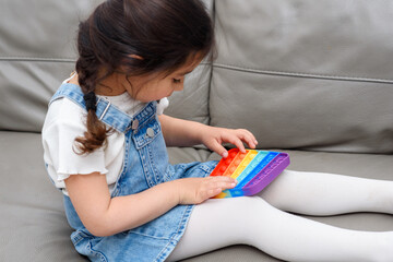 Young Little Girl Sitting On The Sofa At Home Playing Poppit-New Fidget Toy, Popular With Kids,...