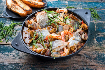Modern style traditional Spanish seafood zarzuela de pescado with fish, king prawns and venus clams served with barbecue garlic bread in red sauce as close-up in design pot