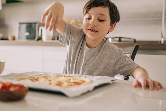Little kid boy making pizza sitting at the table on the kitchen. Children helping in cooking lifestyle image