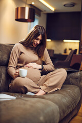 Cute pregnant woman sitting on sofa at home with legs crossed, drinking coffee and cuddling tummy.