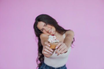 Image of happy smiling woman wearing white dress looking at camera with her finger on her chin and holding piece of cake isolated over pink background
