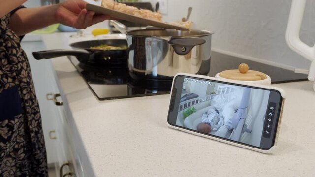 Mother cooking dinner in the kitchen using ip camera as baby video monitor on mobile phone, woman watching her sleeping baby in real-time on smartphone screen. High quality 4k footage