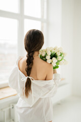 a girl in a white shirt, with a pigtail and a ribbon, stands with her back and holds a bouquet of white tulips