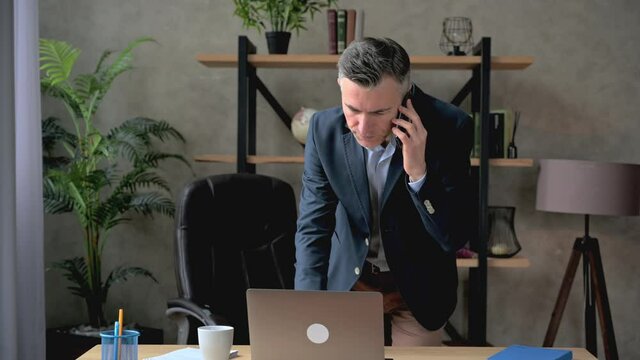 Satisfied busy caucasian successful mature businessman or lawyer talking on the phone with a colleague or customer while using a laptop, browsing the internet, answering an email, smiling
