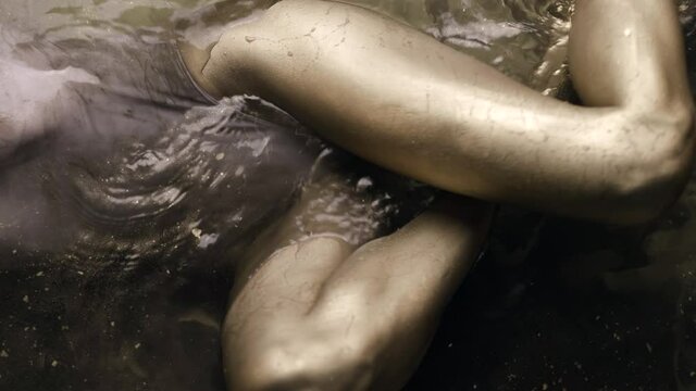 A woman's legs and body in a bodysuit with painted golden skin lie in the water, slowly moving and posing.