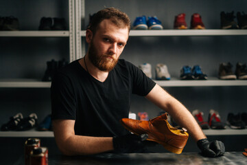 Portrait of handsome bearded shoemaker examining light brown leather shoes during restoration working, looking at camera. Concept of cobbler artisan repairing and restoration work in shoe repair shop.