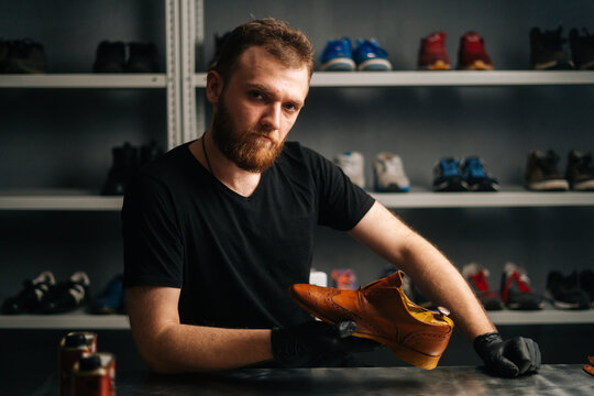 Portrait of handsome bearded shoemaker examining light brown leather shoes during restoration working, looking at camera. Concept of cobbler artisan repairing and restoration work in shoe repair shop.