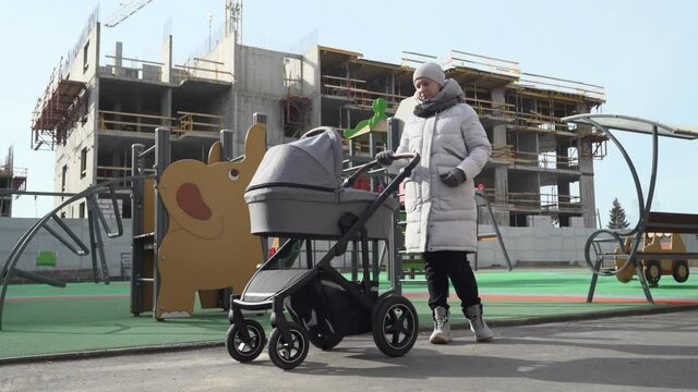 Mother with baby stroller on playground in front of modern apartment building under construction, building of new housing. High quality 4k footage