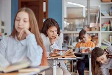 Young girl looks absent-minded in reading. Elementary school kids sitting on desks and reading books in classroom