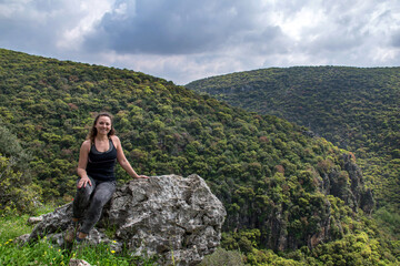 Naklejka na ściany i meble woman traveler sitting on rock and looking at amazing green hills and forest, hiking trip through Israel mountains, wanderlust travel concept, space for text, atmospheric epic moment