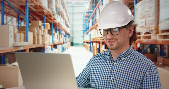 Logistics company manager in white helmet talking on video call using laptop while standing on background of rows with cargo.