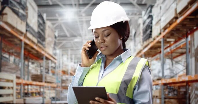 Portrait of young female African American engineer in helmet speaking on smartphone using tablet device standing at construction site. Woman wearing safety equipment at work. Construction concept.