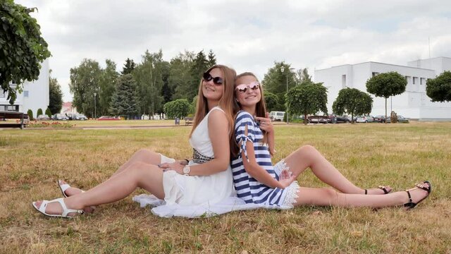 Cheerful female friends in sunglasses are sitting on the grass in the city.