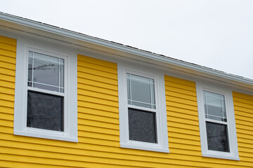 A bright yellow cape cod horizontal siding wood exterior wall with three glass double hung windows and white vinyl eve soffit. The background is a grey cloudy sky.