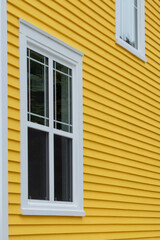 The exterior of a bright yellow narrow wooden horizontal clapboard wall of a house with two vinyl windows. The trim on the glass panes is white in colour. The outside boards are textured pine wood.  