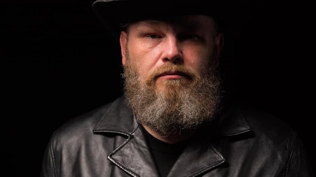 Portrait of a middle aged man with beard in leather coat and an original hat on a black studio background. Man looks away, then turns directly into the camera and tear rolls down his cheek. Close up.