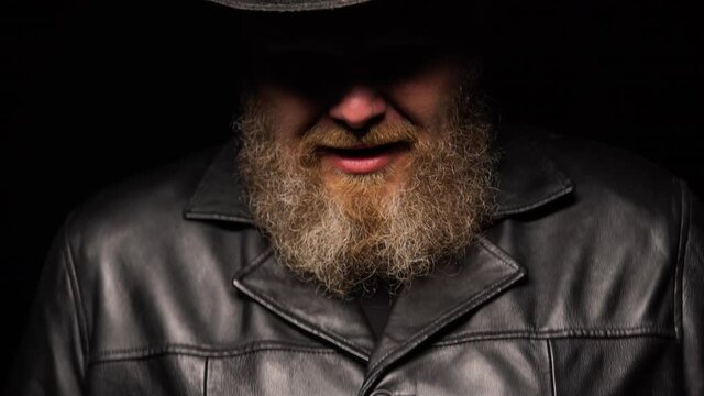 Portrait of a middle aged man with a beard in a leather coat and an original hat on a black studio background. A silhouetted man with a shadow on his face laughs and then screams terribly. Close up.