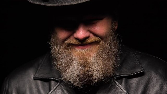 Portrait of a middle aged man with a beard in a leather coat and an original hat on a black studio background. A silhouetted man with a shadow on his face laughs and says kamon baby. Close up.