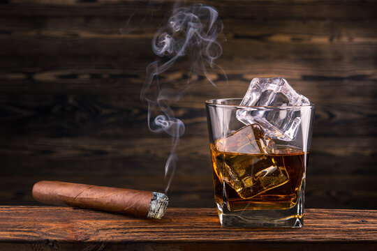 Whiskey and cigar. Glass of whiskey with smoke cigar. Bourbon or Brandy Glass with ice cube and cigars from Cuba Havana on natural wooden background. Alcohol drink on Bar counter in the restaurant. 