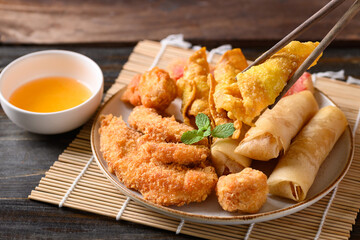 Asian mixed deep fried food (chicken, spring roll, wonton and crab stick) eating with chili sauce by chopsticks on wooden table