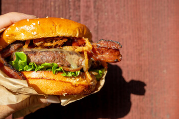 A hand holding a freshly cooked, juicy western BBQ burger with cheese, fried, onions, lettuce, bacon, barbecue sauce on a sesame seed bun in front of a red background.
