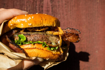 A hand holding a freshly cooked, juicy western BBQ burger with cheese, fried, onions, lettuce, bacon, barbecue sauce on a sesame seed bun in front of a red background.