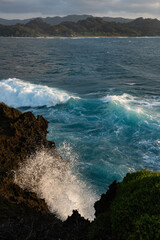 Splash of the sea bursting on the rock with a motion blur, being illuminated by soft sunlight. The choppy emerald blue sea with foams and a mountain in the background on a cloudy day.