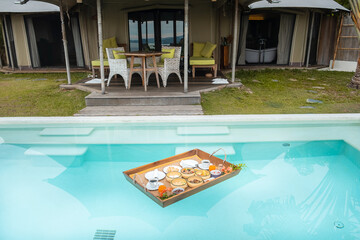 Floating Breakfast tray in swimming pool at luxury hotel or tropical resort villa, fruits, croissant, coffee, and orange juice. Exotic summer, relaxation, tropical travel and vacation concept