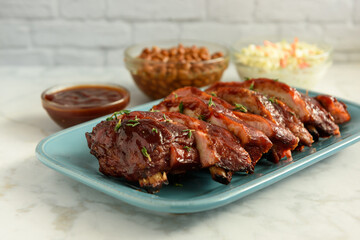 Close up of sliced smoked pork BBQ ribs on a blue plate with sides of BBQ sauce, baked beans and coleslaw on a white marble table and subway tile background