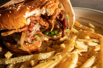 A hamburger with a bite out of it. A freshly cooked, juicy western BBQ burger with cheese, fried, onions, lettuce, bacon, barbecue sauce on a sesame seed bun on a plate piled with garlic french fries.