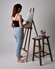 Full length portrait of a red haired artist girl wearing casual jeans and tank top. standing pose painting a canvas on an easel, against a studio background.