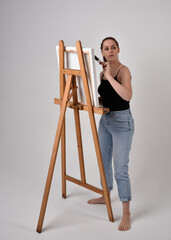 Full length portrait of a red haired artist girl wearing casual jeans and tank top.  standing pose 
painting a canvas on an easel, against a studio background.