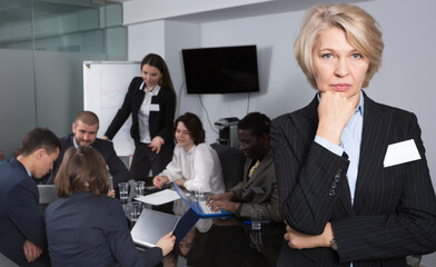 Unhappy mature business woman standing in meeting room on background with coworkers.