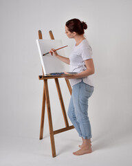 Full length portrait of a red haired artist girl wearing casual jeans and white shirt.  standing pose 
painting a canvas on an easel, against a studio background.