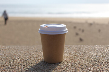 A cup of Coffe, is on the surface of a small rock, in the background a man on the beach and pigeons, blue sky