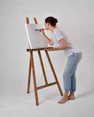 Full length portrait of a red haired artist girl wearing casual jeans and white shirt.  standing pose 
painting a canvas on an easel, against a studio background.