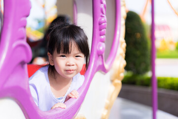 Cute Asian girl sitting in carousel ride. Happy child sit and watch from window of the player. Kid...