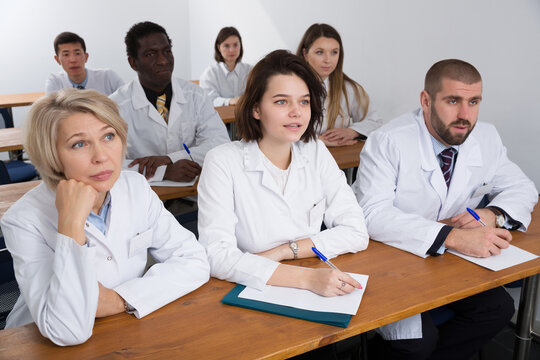 Multinational positive group of people in white coats attentively listening and making notes while sitting in boardroom