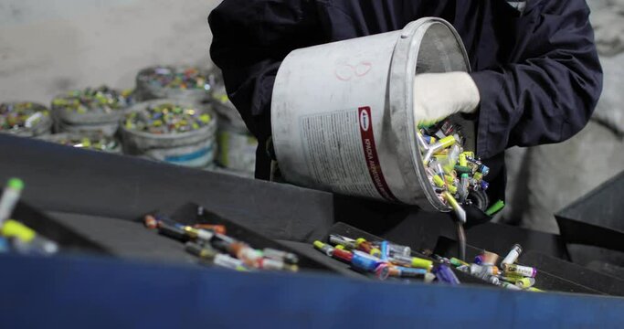 View of hands of person in uniform and gloves pouring out used batteries from bucket onto a moving conveyor belt, for separation and recycling. Processing. Environmental protection concept.