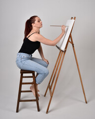Full length portrait of a red haired artist girl wearing casual jeans and white shirt. Sitting pose on chair, painting a canvas on an easel against a studio background