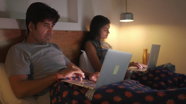 A young attractive hard-working  and tiring woman and man are sitting on a bed and working late in the night on an office project or assignment using their respective. laptops. work from home concept.