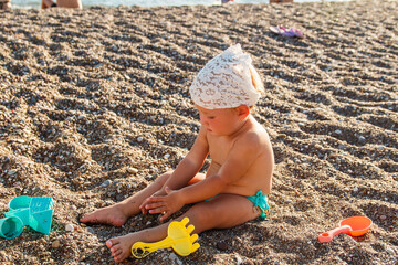 A cute little girl in bathing trunks sits on the sand on the beach and plays with sand shovel. Tanned baby in the summer