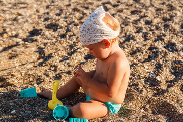 A cute little girl in bathing trunks sits on the sand on the beach and plays with sand shovel. Tanned baby in the summer
