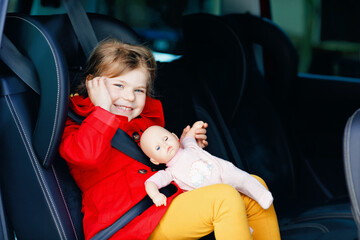 Toddler girl sitting in car seat, holding favourite doll toy and looking out of the window on nature and traffic. Little kid traveling by car. Child safety on the road. Family trip and vacations