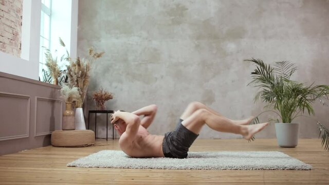 A young european man works out on the background of the grey wall, doing abdominal exercises on a wooden floor while lying on a mat. Side view. Healthy lifestyle and sport concept.