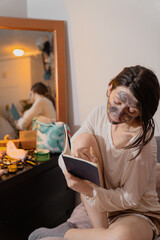 Woman doing black charcoal clay face cleansing mask at home in pajama, enjoying skincare ritual. Candid photo