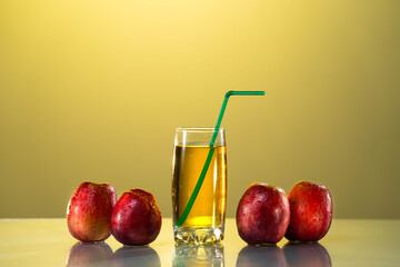 Red apples lie on the table with a reflection. next to it is a glass of apple juice and a straw. sunset background. yellow background. Glass of apple juice on blue background